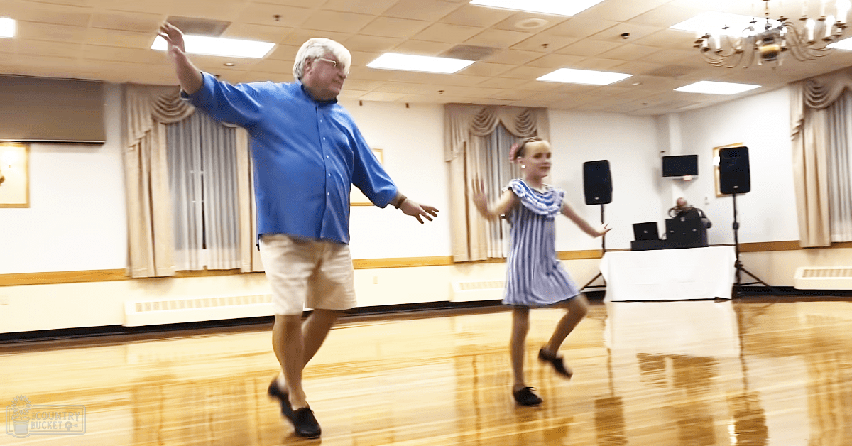 An adult male dancing with a young female in a dance studio, surrounded by wooden floors, curtains, speakers, a chandelier, and a TV on the wall.