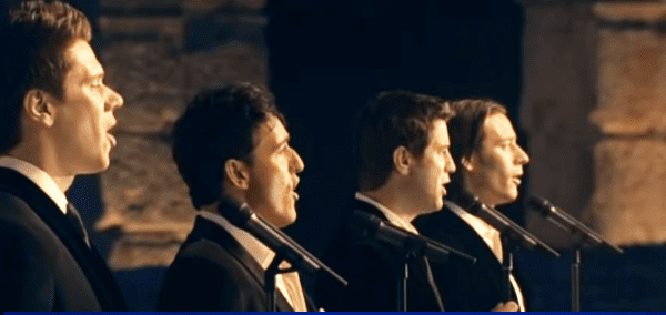 A male choir performing indoors under dim evening light against a backdrop of stone columns.