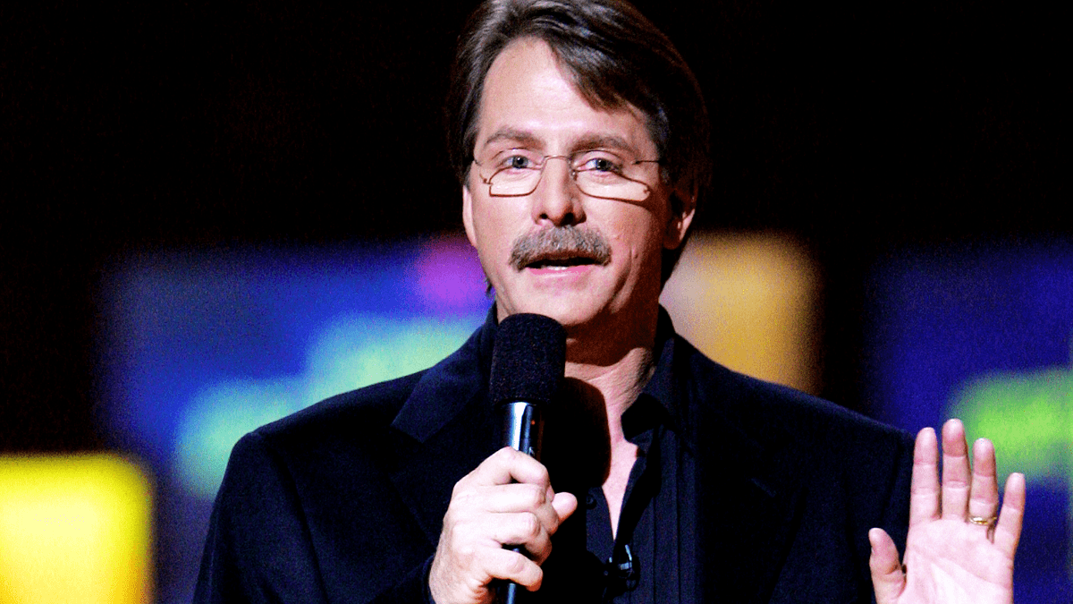 A middle-aged man wearing a dark blazer and shirt speaks into a microphone at an indoor event space, likely during an evening performance or conference.