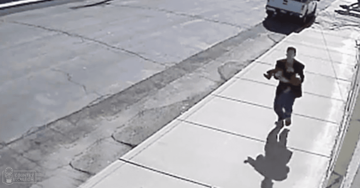 An adult male holding an infant in a light-colored outfit outdoors in a parking lot with concrete tiles and shadows indicating late afternoon.
