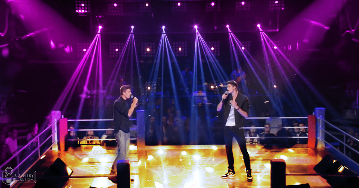 Young adult male performers in a theater or concert hall with stage lighting and an audience behind barriers.
