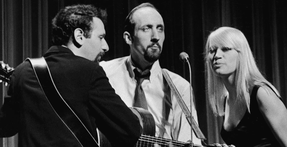 A young adult male musician in a dark-colored formal suit plays guitar on an indoor stage with two other individuals visible in the background, one wearing a light-colored suit jacket and another in a black sleeveless top.