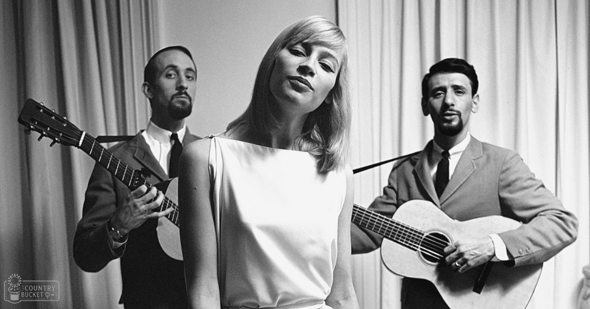 A woman in a white sleeveless top stands among two men dressed formally on what appears to be a stage with vertical curtains, suggesting an indoor performance venue.