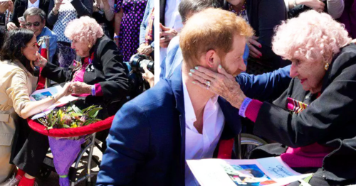 An adult male singer interacts with an older adult female holding a bouquet of flowers, set against a backdrop of a community event on a sunny day.