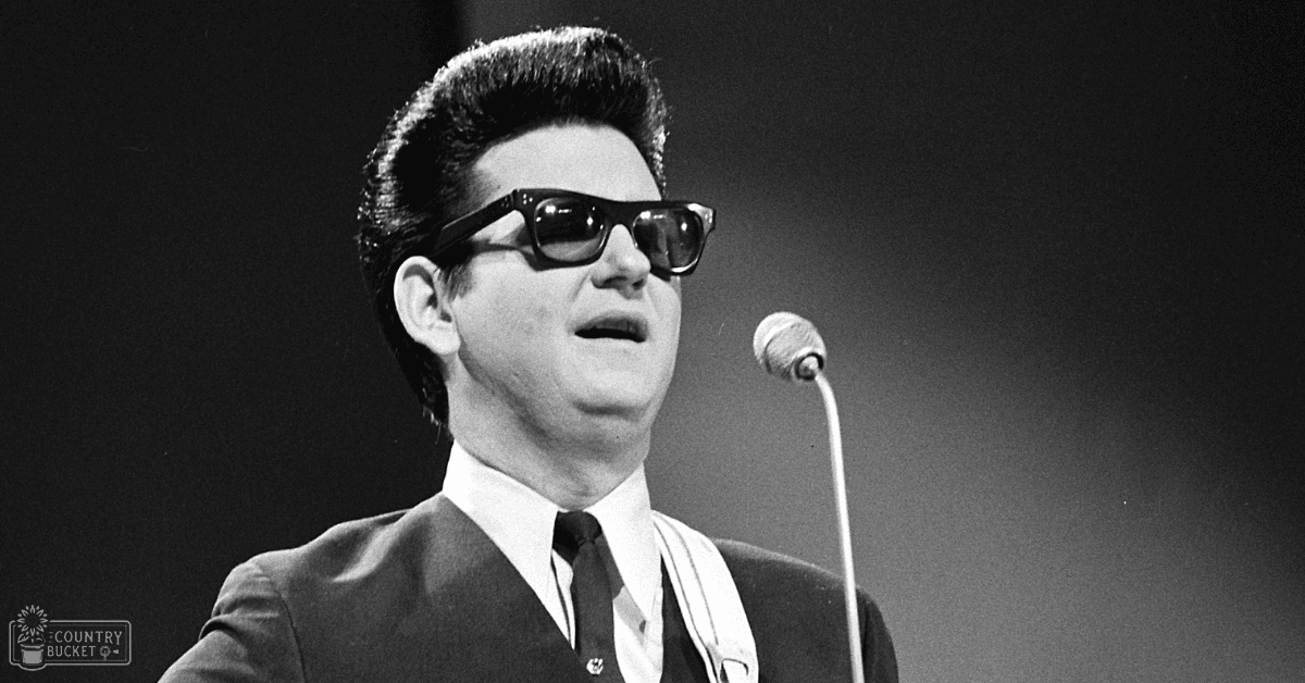 A male performer in a dark suit and white shirt on stage, likely from the mid-20th century, with a microphone to his right under dimmed lighting.