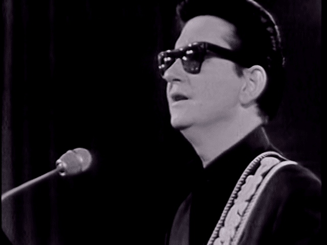 An adult male performer on stage with a dark-patterned jacket, microphone prominently featured, dramatic lighting typical of live performances.