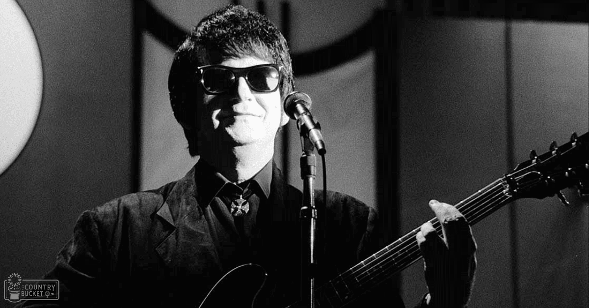 A male adult musician wearing a dark suit jacket with a notched lapel and sunglasses performs on stage with an acoustic guitar under professional lighting.