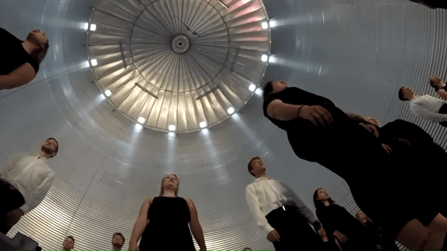 Adults in an indoor setting with a large fan-like structure overhead, wearing black and white tops under artificial lighting.