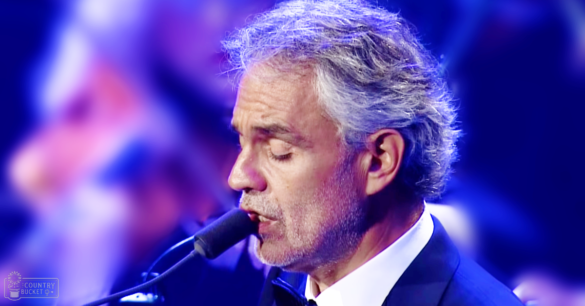 A middle-aged man sings into a microphone, dressed in a tailored dark blue suit with a white collared shirt, at what appears to be an indoor concert or performance setup.