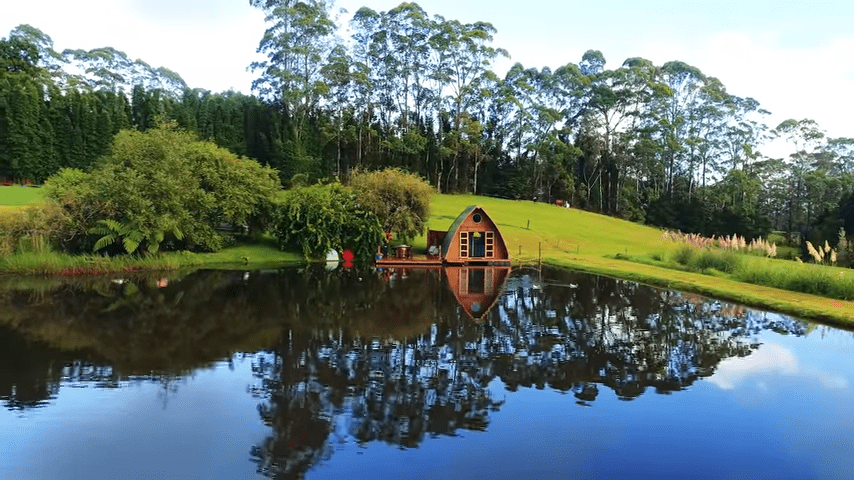 A small wooden A-frame cabin on stilts over calm waters in a serene natural setting with lush greenery and vibrant spring or summer foliage.