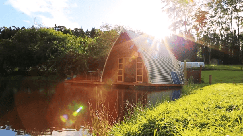 A serene outdoor scene with a small wooden cabin with solar panels near water, lush greenery, and a body of water reflecting sunlight.