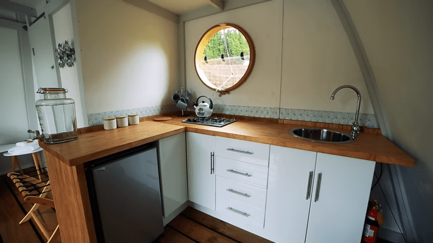 A cozy indoor kitchen scene with a round window showing greenery, white cabinets, a wooden countertop, stainless steel sink, small refrigerator, glass jar, ceramic containers, kettle, and other utensils.