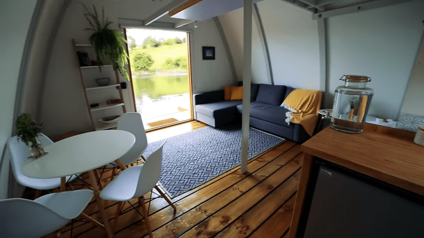 A serene, modern interior with a white oval dining table, dark blue sectional sofa adorned with yellow throw pillows and blanket, patterned rug, wooden countertop, bookshelf against the wall, large window showing greenery outside. Daytime natural sunlight streaming in.