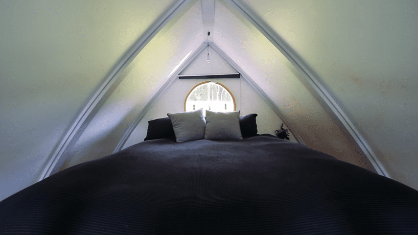 An attic bedroom with sloped ceilings, dark bedding, white walls, natural light through a small circular window, and decorative items in the corner.