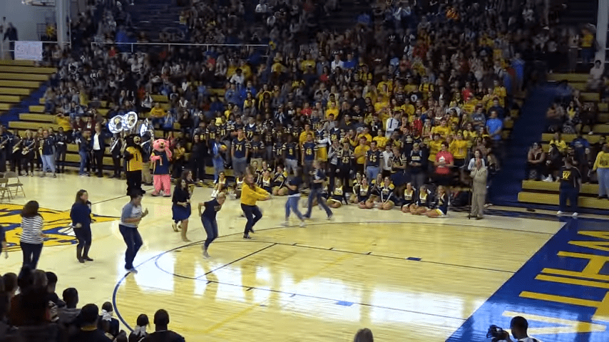 A dance performance by children and young adults in a brightly lit indoor sports arena with bleachers filled with spectators wearing casual school spirit attire. The season appears to be fall or winter, based on the clothing worn.