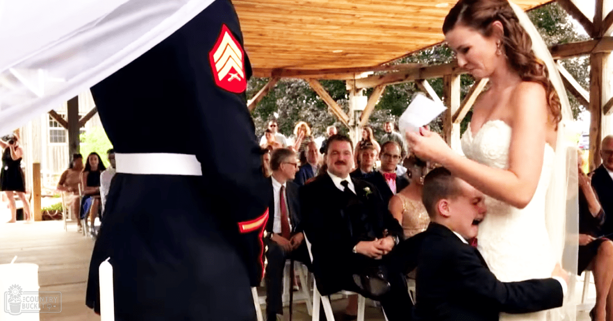 An adult female bride in a white lace wedding gown with a veil, kneeling during an outdoor wedding ceremony at a wooden pavilion with seated adults and a groom in a dark military uniform with red insignia.