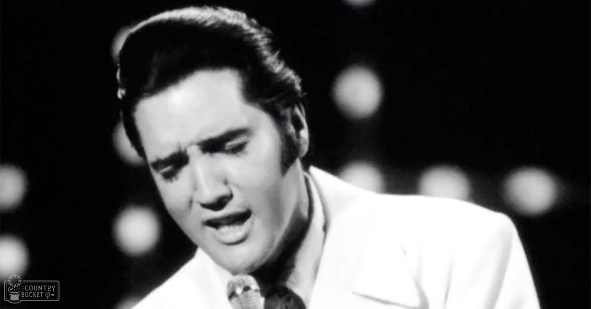Adult male performer wearing white suit jacket and dark tie, possibly from mid-20th century, in an indoor performance venue with stage lighting.