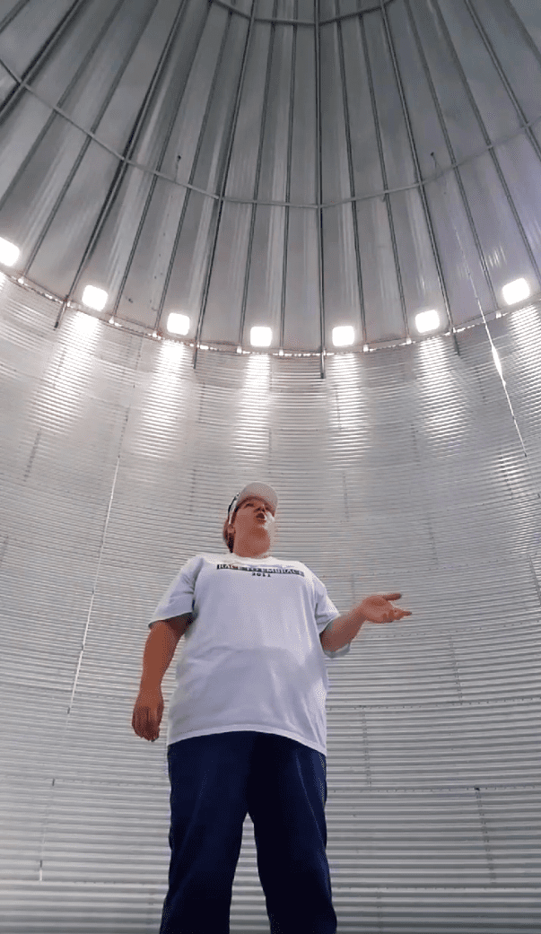 Young adult male in a relaxed, casual white t-shirt with 'FLORIDA 2018' text and dark blue pants inside an industrial silo or storage structure.
