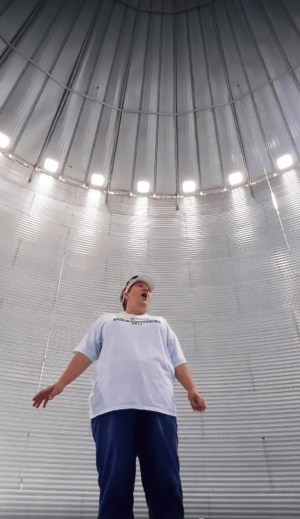 A young adult male stands inside an industrial silo with corrugated metal walls, wearing a white T-shirt and a white cap with text on it.