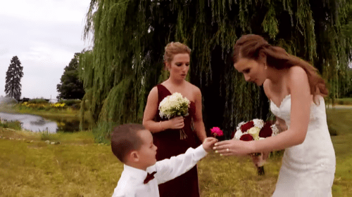 A young male toddler wearing a white shirt with a dark red bow tie interacts with two adult females in a wedding setting outdoors, surrounded by a willow tree and a body of water.