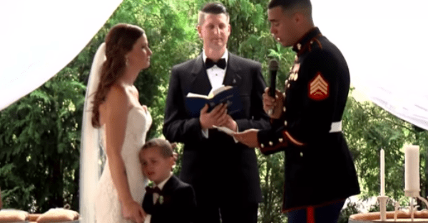 A wedding ceremony outdoors under a white canopy with greenery surrounding it. The bride wears a white gown and veil; groom is dressed in a black tuxedo, officiant holds a microphone and book, child stands between them wearing a suit jacket.