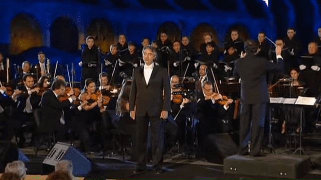 A conductor leading an orchestra in a concert setting with various ages of adult musicians and dark blue backdrop.