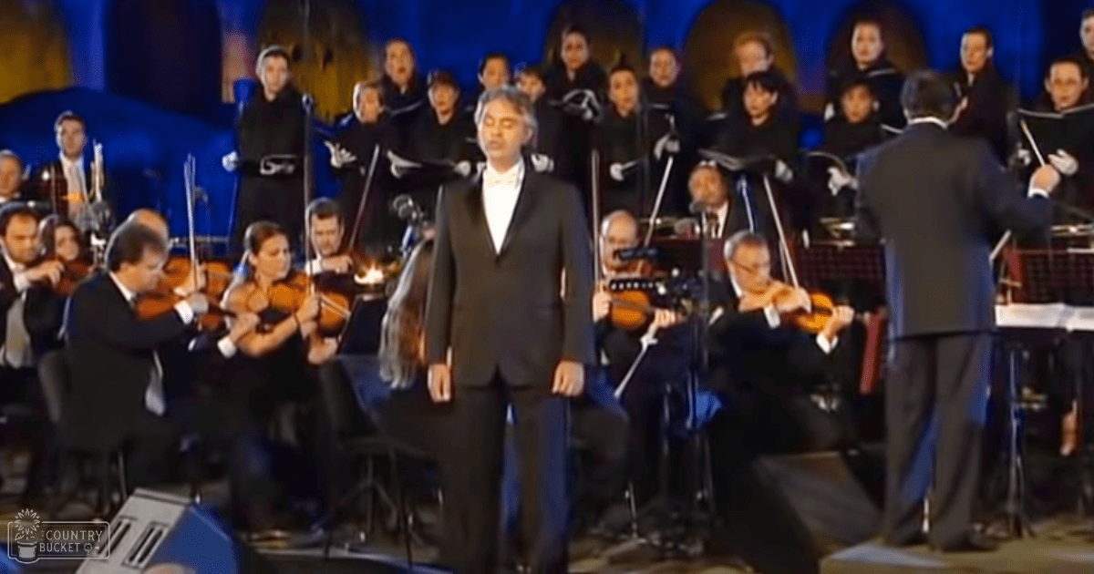 Adult male singer performing with an orchestra and choir in a concert hall setting, dressed in formal attire under blue stage lighting.