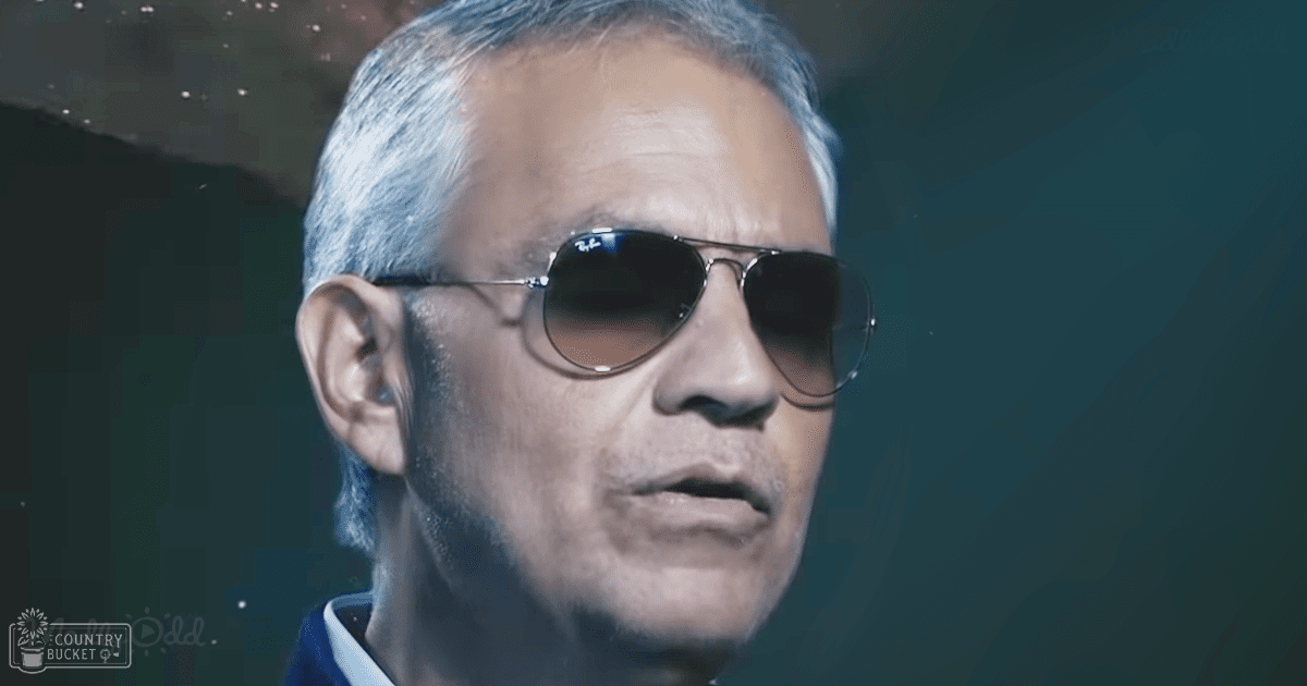 An older adult male in a dark setting wearing aviator-style sunglasses with 'Madly Odd' text on his garment, possibly indoors with dim lighting and some light reflections.