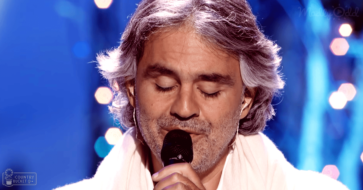 A middle-aged man in a light-colored tunic sings into a black microphone under blue stage lights at an indoor performance venue.
