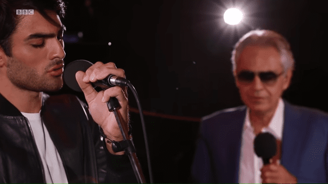 A young adult male singer with a microphone, accompanied by an older adult male wearing sunglasses standing nearby holding another microphone; the scene takes place indoors during what seems to be a music event or recording session at night.