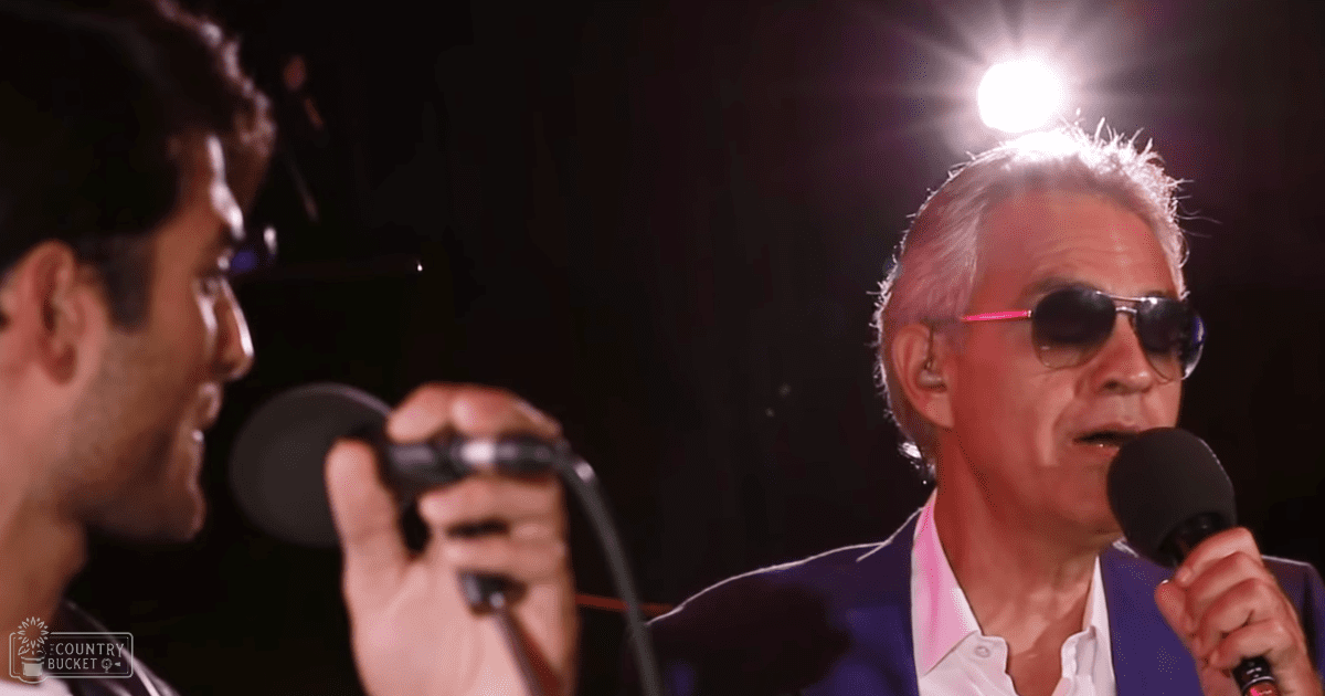 A middle-aged man singing into a microphone, wearing a blue suit over a white shirt with dark sunglasses, performing under stage lighting in an indoor concert setting.