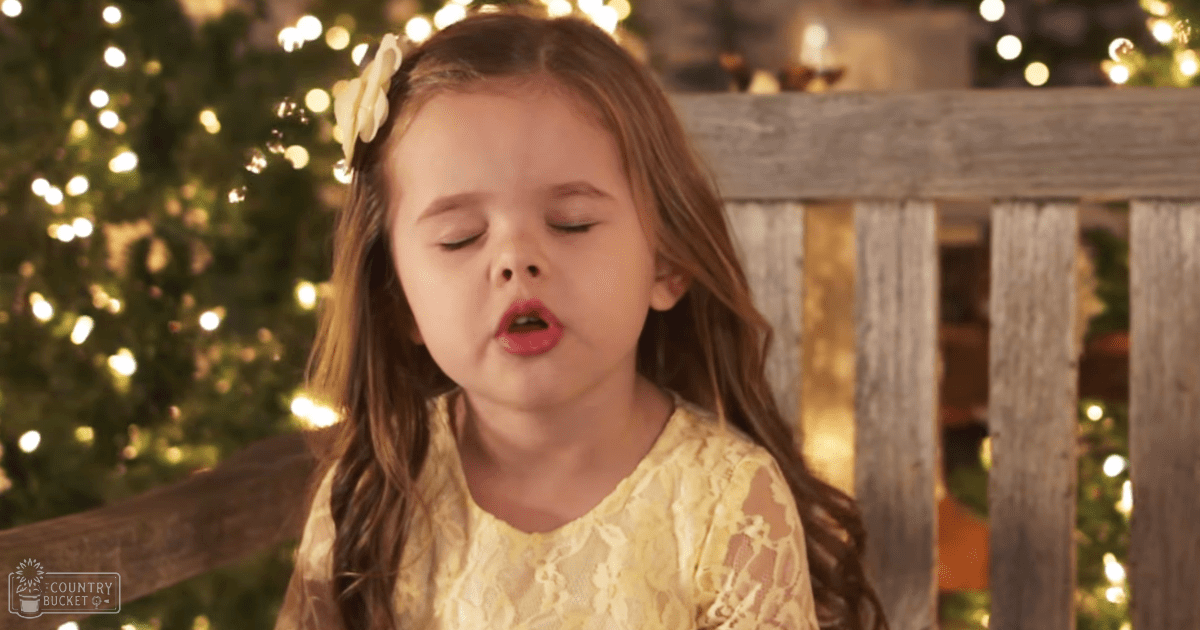 A toddler girl in a bright yellow lace dress with floral patterns, standing indoors amidst festive decorations.