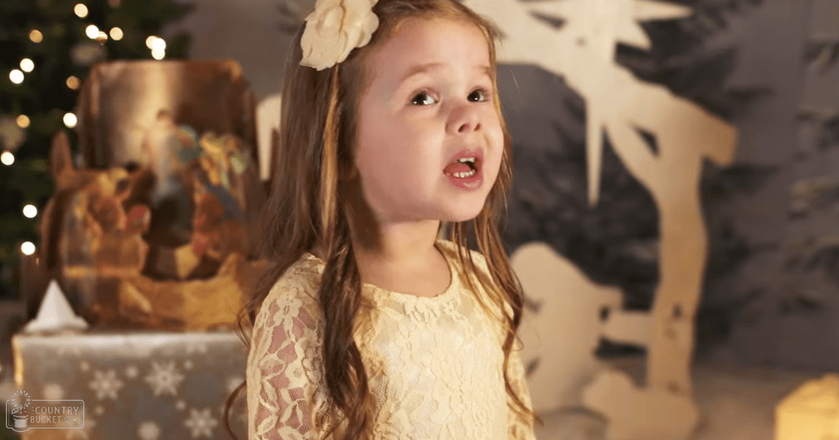 A toddler in a festive indoor setting, wearing a cream lace dress with long sleeves and floral patterns, surrounded by Christmas decorations including a reindeer and presents.