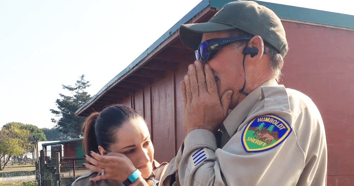 A man in a tan sheriff's uniform with a badge reading "HUMBOLDT SHERIFF" stands outside near a red building with a green roof, surrounded by trees and fencing under bright daylight.