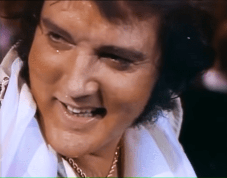 A middle-aged adult male wearing a loose, white traditional garment with intricate patterns stands indoors under bright stage lighting.