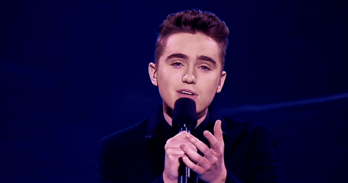 A young adult male in a dark blazer performing on stage with focused lighting and a microphone.