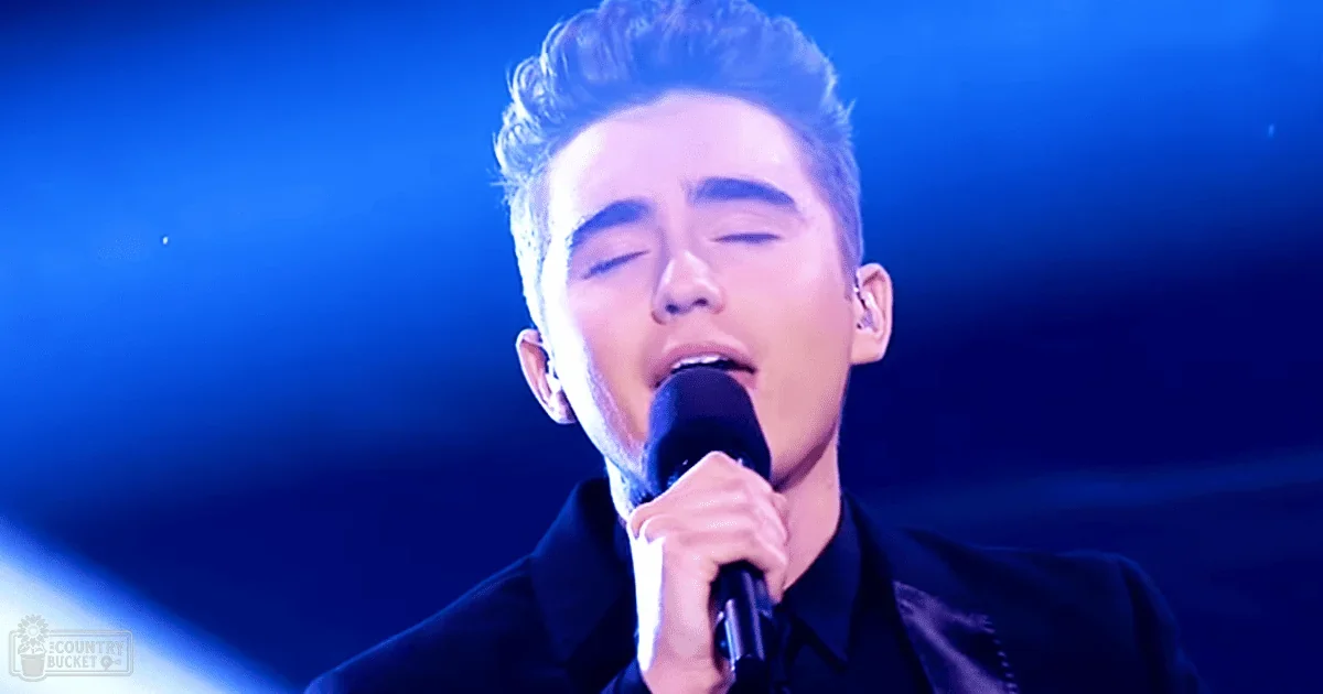 A young adult male singer performing on a stage with dramatic blue lighting, wearing a fitted black shirt.