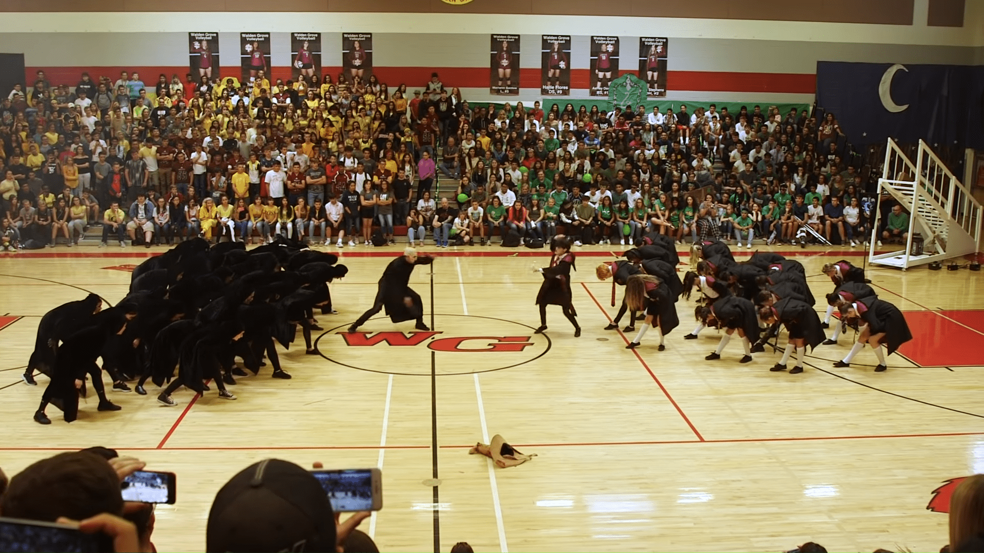 A dance performance at a school gymnasium with young adults in black attire, an audience of children and teenagers seated on bleachers, and a logo reading 'WG' on the floor.