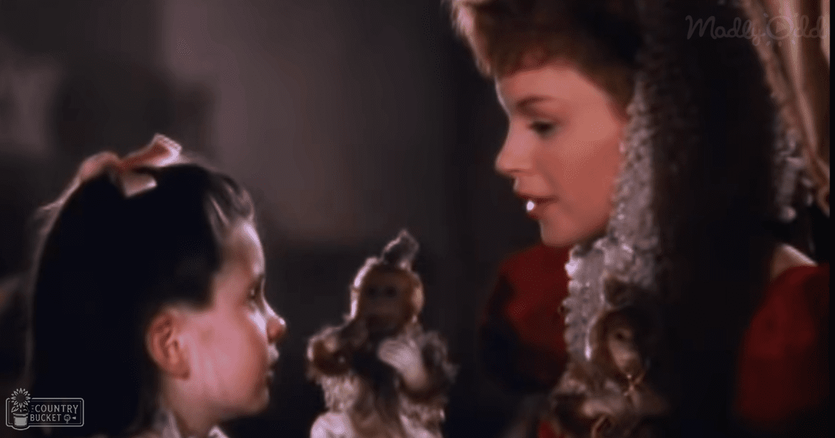 A young girl dressed in a dark-colored historical-style dress stands next to an adult woman wearing a richly detailed Victorian-era red garment, both in an indoor setting with dim lighting.