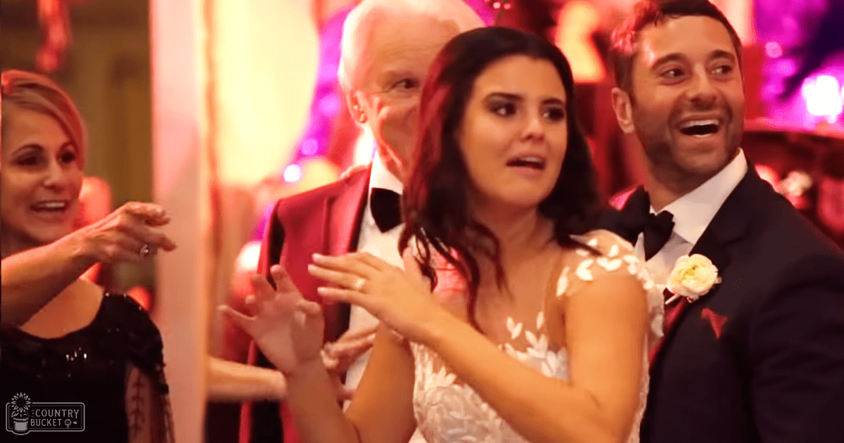 A wedding scene indoors, with a bride in a white floral-patterned gown with a small flower accessory and an older man in a red suit jacket over a white shirt with a black bow tie. The setting includes blurred lights suggesting evening or night time.