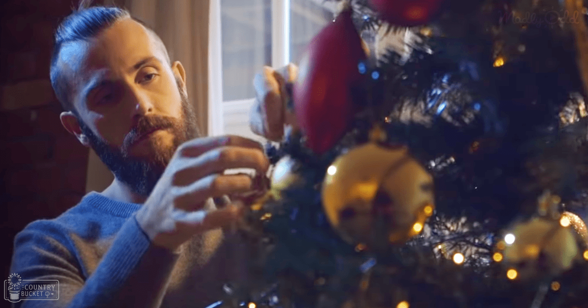An adult male decorates a Christmas tree indoors under artificial lighting, wearing a blue long-sleeved sweater with a subtle pattern in a cozy winter evening setting.