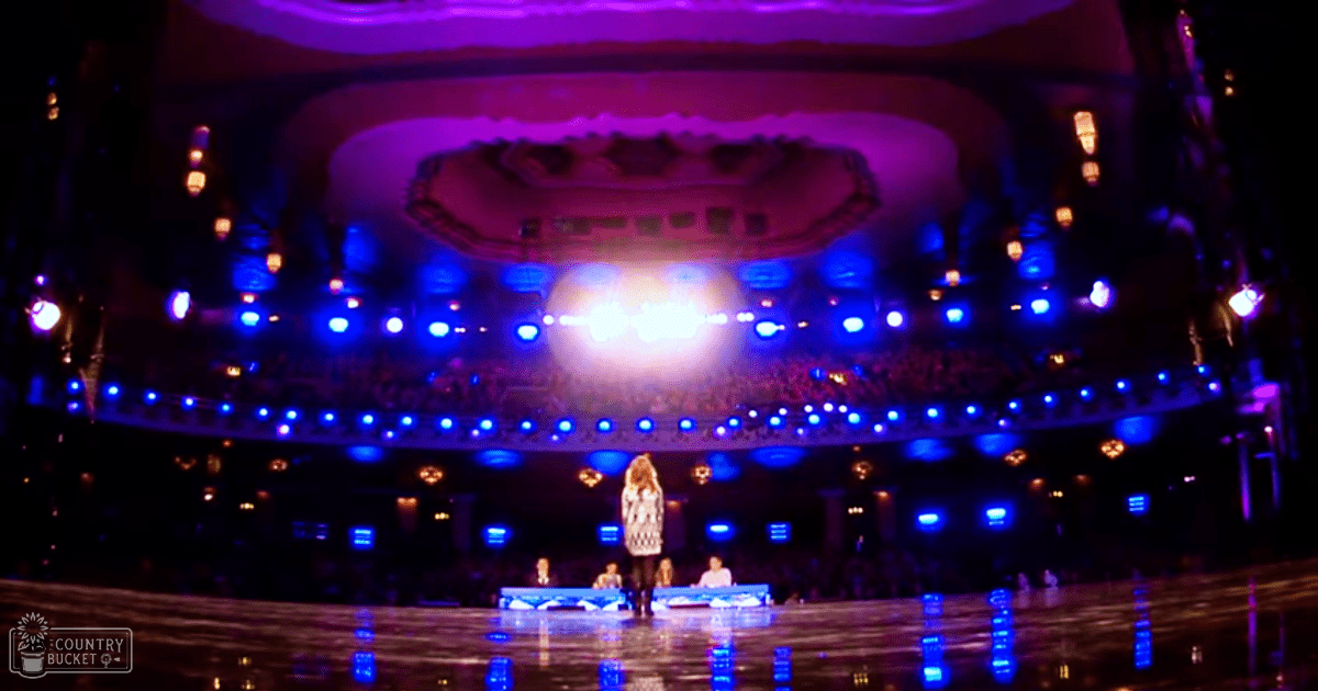 An adult female singer performs on a stage in an ornate indoor theater with bright chandeliers and reflective flooring.