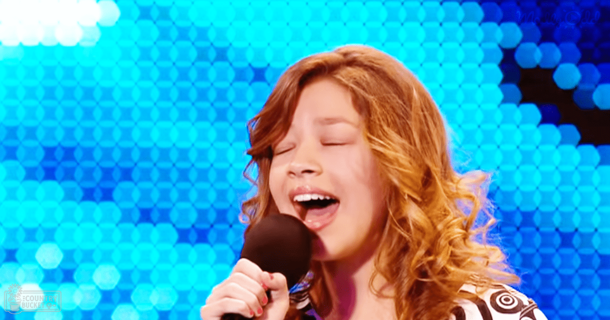 A young adult female sings into a microphone, dressed in a patterned V-neck top with short sleeves and light fabric, against a blue hexagonal stage backdrop.