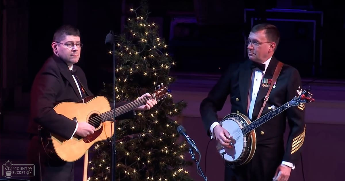 A middle-aged male singer in a black suit performs alongside another male performer in a military-style uniform with gold epaulets, both on stage near a Christmas tree during what appears to be a holiday concert or celebration.