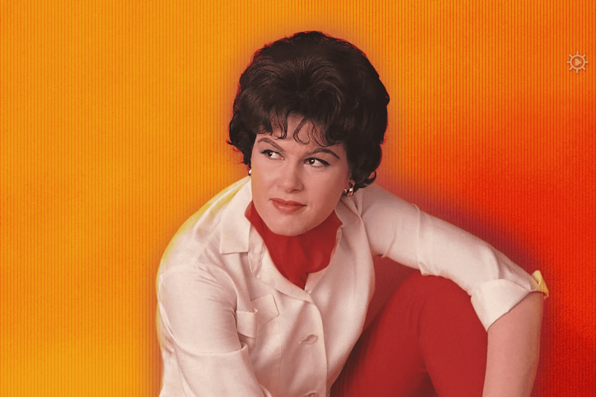 A middle-aged adult female wearing a white blouse with red turtleneck and pants, possibly from the 1960s, in an indoor studio setting with bright orange lighting.