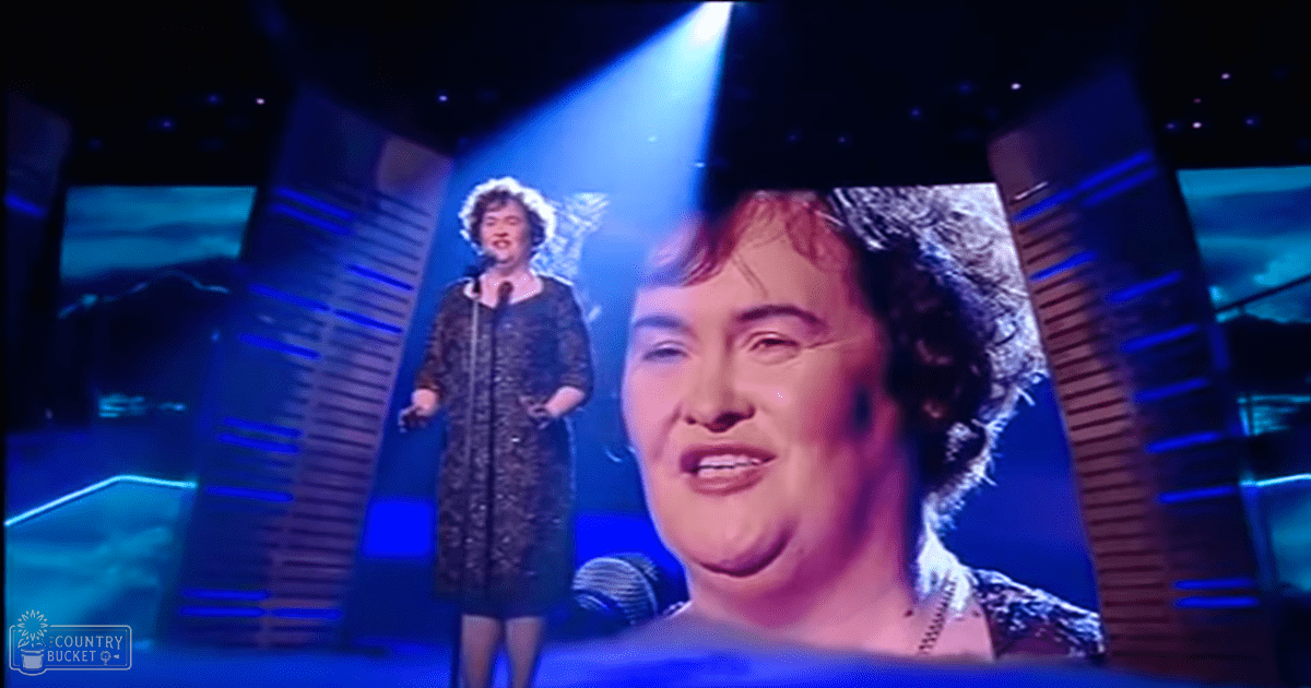 A female adult singer in a dark patterned dress performing on an indoor stage with blue lighting, holding a microphone.