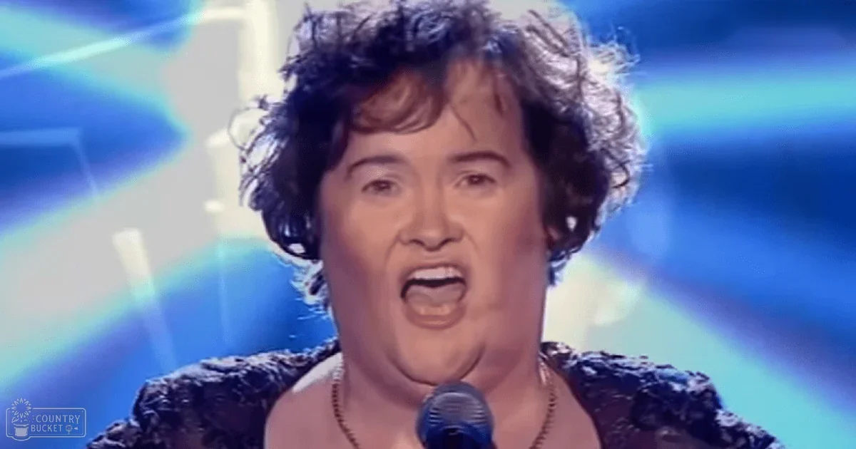 A middle-aged female singer in a talent show, wearing a dark patterned top with lace-like details and short sleeves, performing on stage under bright blue lighting.