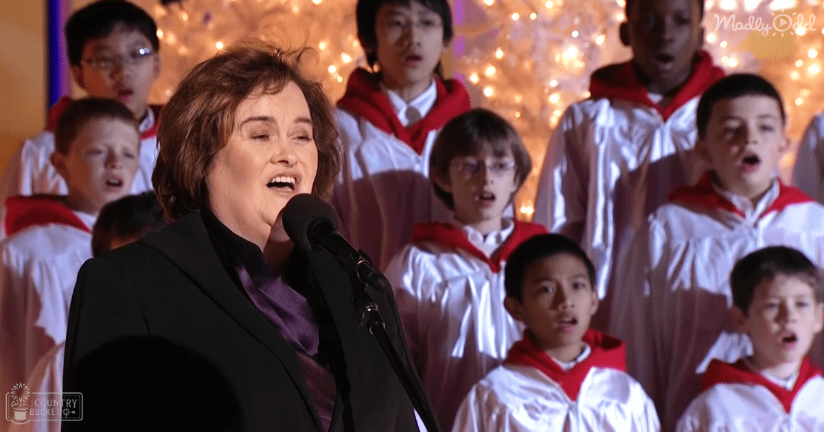 A middle-aged to older adult female sings in front of a choir dressed in white robes with red trim on what appears to be an indoor stage decorated for Christmas.