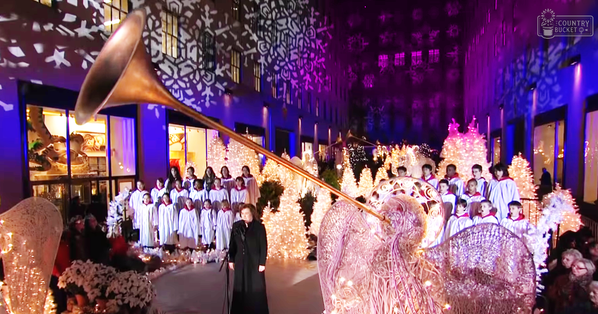 A Christmas carol performance with children in white robes and a conductor, set against festive decorations inside an indoor shopping mall during the evening.