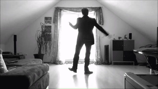An adult male in an attic room wearing a dark suit jacket and dress shoes, surrounded by furniture and decor with natural light streaming through the window.
